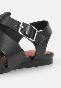 Black leather sandal with crisscross straps, silver buckle detail, and a flat black sole. Brown insole is partially visible.