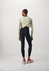 Šviesiai žalios spalvos ilgomis rankovėmis iškirptas cropped topas su kryžminiu nugaros dizainu, derinamas su juodais leggingais ir rožiniais sportiniais batais. Lygi tekstūra, prigludęs dizainas.