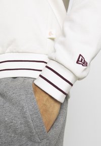 Felpa bianca con righe bordeaux ai polsini e un piccolo tag con logo. Abbinata a pantaloni grigi chiari con dettagli sulle tasche.