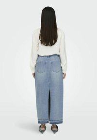 Jupe maxi en denim bleu clair avec une fente haute et un design à cinq poches. Le modèle porte une chemise blanche à manches longues et des escarpins noirs.