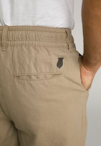 Pantalons légers beiges avec une taille élastique, poches latérales et une poche arrière avec un logo en forme de bouclier foncé. Texture de tissu lisse.