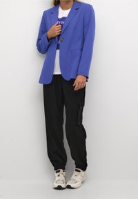 Blazer su misura blu con un solo bottone, abbinato a una t-shirt bianca con grafica e pantaloni cargo neri con polsini elastici. Sneakers bianche completano il look.