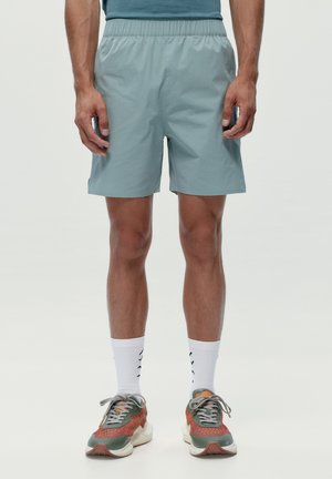 Uomo con pantaloncini azzurri, calzini bianchi fino a metà polpaccio e scarpe da ginnastica grigie, arancioni e verdi, in piedi su uno sfondo semplice.