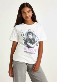 Valkoinen puuvillainen t-paita, jossa on hahmon kuva, jolla on tumma hiukset. Teksti "WEDNESDAY DESTINED FOR DARKNESS" violettina. Lyhyet hihat.