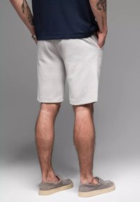 Hellgraue Baumwollshorts mit lockerer Passform, seitlichen Taschen und einem Saum, der bis zur Mitte des Oberschenkels reicht. Getragen mit grauen Slippern.