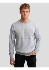 Mand med blondt hår iført en lysegrå sweatshirt med teksten "BIG STAR Your Life Your Jeans", stående med den ene hånd i lommen.