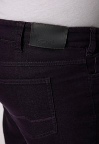 Pantalons en denim violet foncé avec une finition texturée, dotés d'un patch en cuir gris portant l'inscription "MEN+" et de détails de couture visibles.