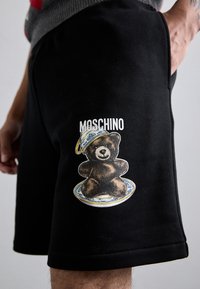 Fekete pamut rövidnadrág, amelyen egy sapkás medve grafika és tányér minta látható, a "Moschino" márkanevével fehér színben.