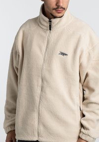 Giacca in pile beige con colletto alto, chiusura con zip e tessuto strutturato. Presenta un piccolo logo e tasche laterali. Vestibilità rilassata.