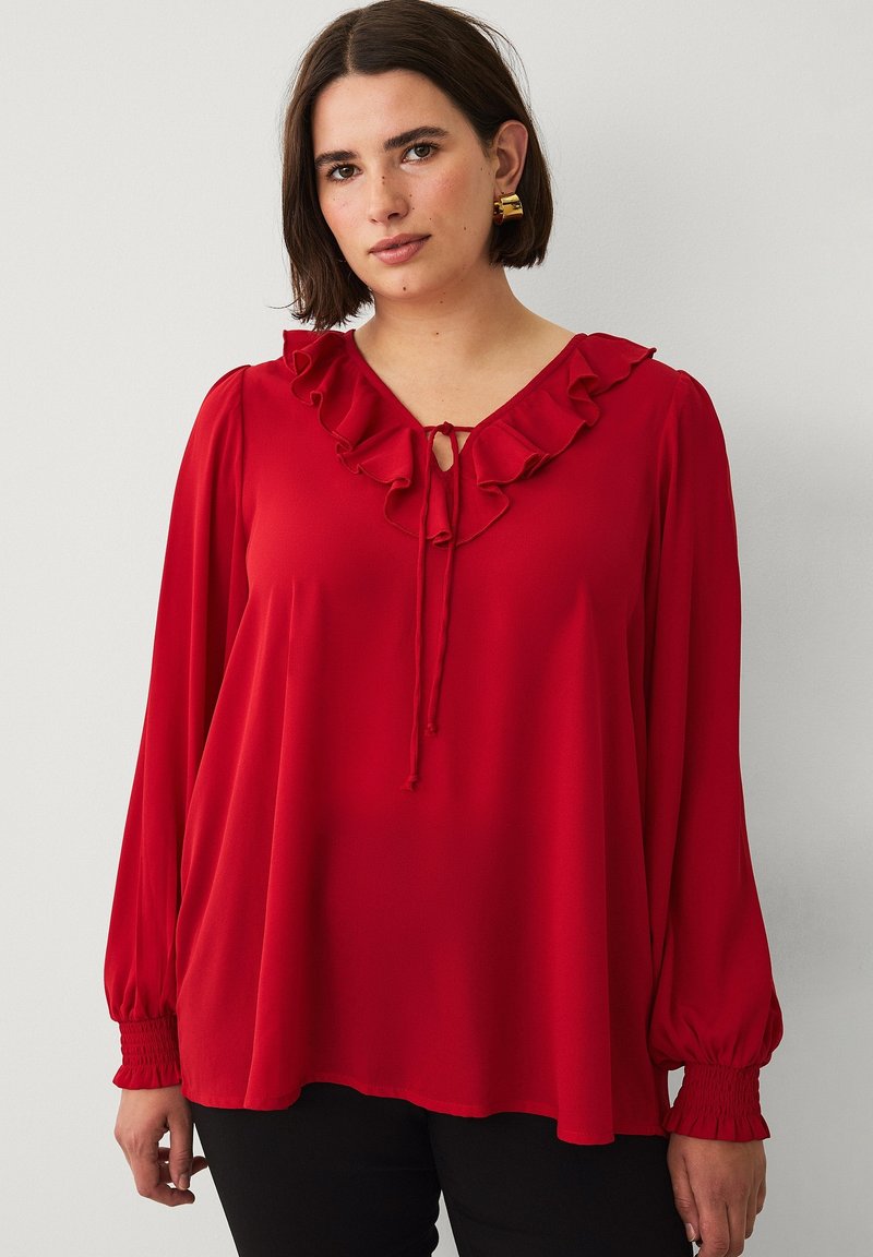 Rode blouse met ruches bij de hals, een sleutelgaatje en lange mouwen. Gemaakt van een gladde stof met gathered manchet. Gstyled met zwarte broek.