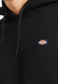 Felpa nera con cappuccio realizzata in tessuto morbido, dotata di tasca a marsupio, cappuccio con cordoncino e una patch rettangolare con il logo Dickies sul petto.
