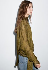 Gennemsigtig olivengrøn top med høj hals, lange ærmer og slangeskindsprint, parret med lyseblå denimjeans.