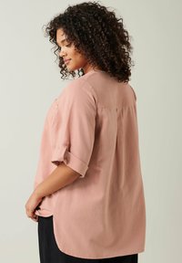 Vaaleanpunainen, kevyt paita rennolla istualla, käännetyillä hihoilla, nappiyksityiskohdalla, takapuolella halkiolla ja pidemmällä helmalla takana lisäsuojan vuoksi.