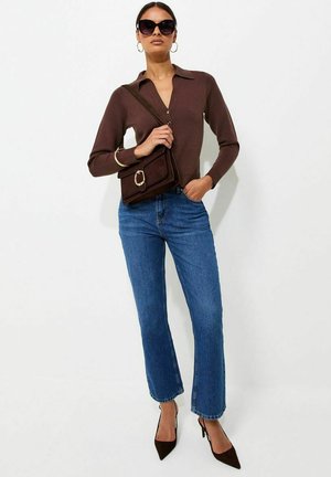 Donna che indossa una maglietta a maniche lunghe marrone scuro, jeans blu, tacchi neri, grandi occhiali da sole, orecchini a cerchio dorati e porta una borsa a spalla marrone abbinata.