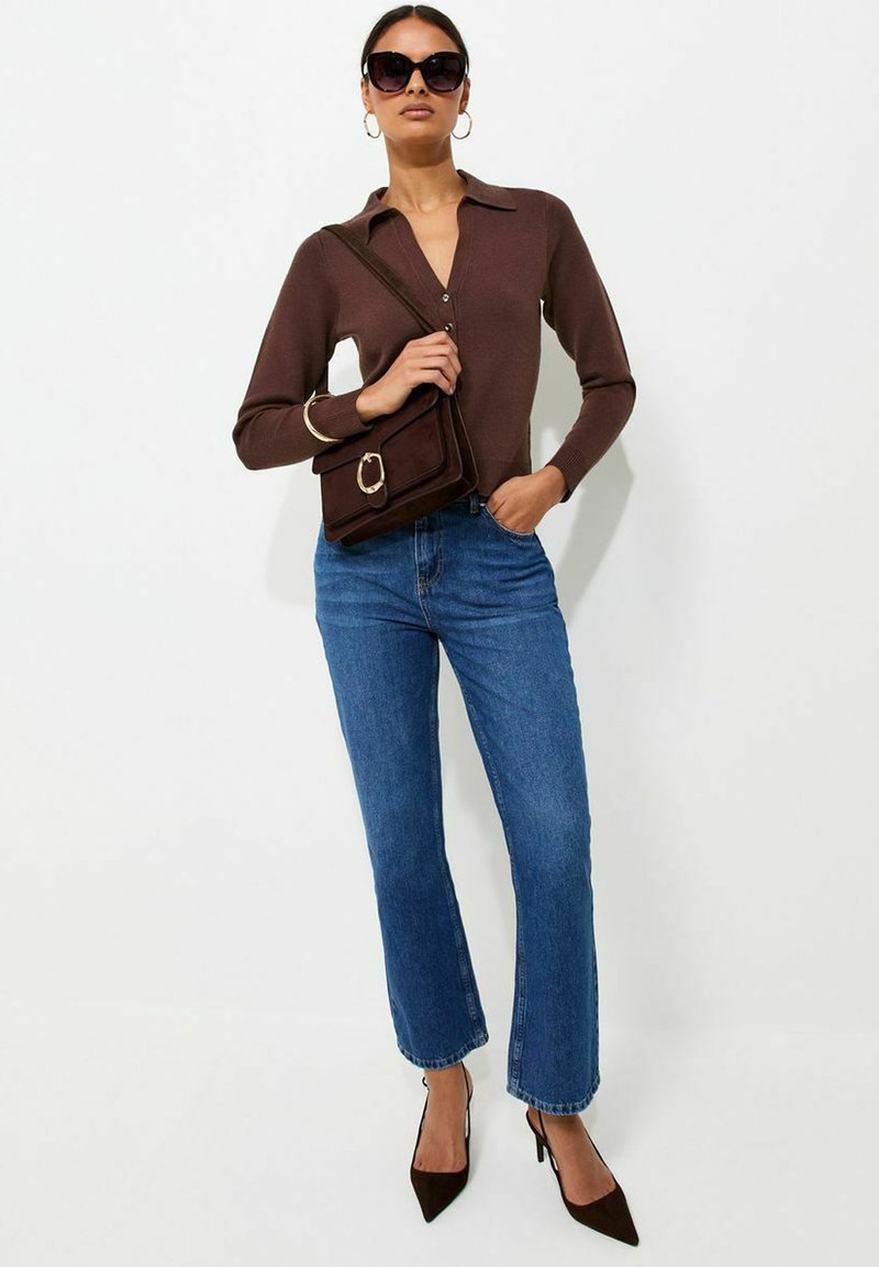Donna che indossa una maglietta a maniche lunghe marrone scuro, jeans blu, tacchi neri, grandi occhiali da sole, orecchini a cerchio dorati e porta una borsa a spalla marrone abbinata.