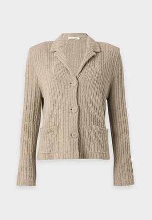 Femme portant un blazer en maille beige, une chemise noire, un jean bleu à jambes larges, tenant un sac à épaule noir, debout devant un fond uni.