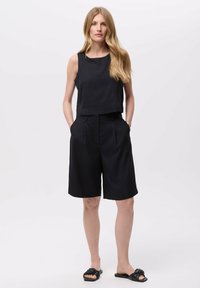 Zwarte cropped tanktop gecombineerd met hoge zwarte short met plooien. Model draagt zwarte slidesandalen en heeft steil haar.