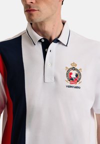Polo en blanco con detalles en azul marino y rojo. Presenta un cuello clásico, tres botones y un logo bordado en el pecho. Tejido texturizado.