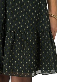 Robe en tissu vert foncé avec petits motifs paisley jaunes et marron, ourlet à volants et bracelet chaîne doré au poignet.