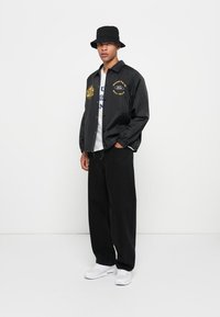 Black windbreaker jacket with gold embroidery, gray t-shirt, loose black pants, and white sneakers. Accessories include a black bucket hat.