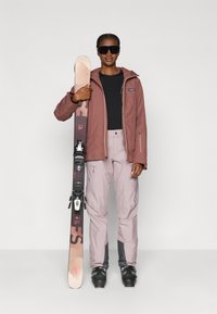 Sífelszerelés, amely porrózsaszín vízálló dzsekit, fekete aláöltözetet, világos rózsaszín sínadrágot és fekete sícipőt tartalmaz. Sílécek kötésekkel a kezében.