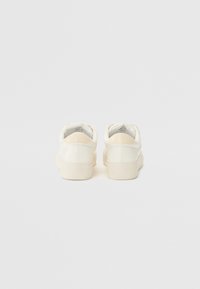 Baskets en cuir blanc avec une texture lisse, présentant des bords arrondis et un accent beige sur le talon, sur un fond uni.
