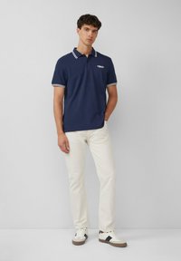Navyblaues Poloshirt mit weißen Akzenten und kurzen Ärmeln, kombiniert mit cremefarbenen Hosen und weißen Sneakers. Model steht vor einem hellen Hintergrund.