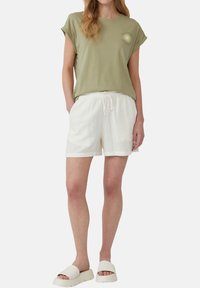 Olijfgroene katoenen T-shirt met opgerolde mouwen en zonafbeelding; witte textuur shorts met koord in de taille; lichte sandalen met dikke zool.