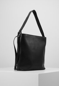 Borsa a spalla in pelle nera con ampia tracolla e ulteriore tracolla sottile regolabile, esposta su un piedistallo bianco contro uno sfondo neutro.