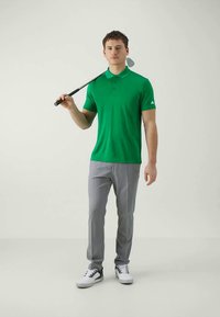 Tânăr îmbrăcat într-o bluză polo verde și pantaloni gri, ținând un caddy de golf sprijinit pe umăr, stând pe un fundal neutru.