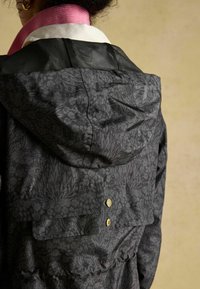 Čierna bunda s kvetinovým vzorom a kapucňou; má textúrovanú tkaninu a viditeľné zlaté kovové akcenty. Rúžová golier dodáva detail.