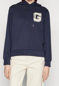 Felpa con cappuccio blu navy in tessuto morbido, caratterizzata da un grande logo "GANT" di colore crema e cordini sul cappuccio. Taglio rilassato.