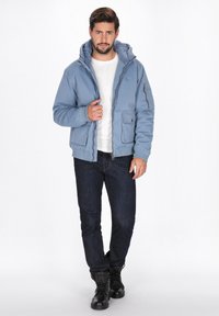 Lysblå dunjakke med hætte, frontlynlås og sidelommer, båret over en hvid t-shirt og parret med mørke jeans og sorte støvler.