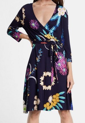 Femme portant une robe portefeuille bleu foncé à motif floral, manches trois-quarts et ceinture nouée à la taille.