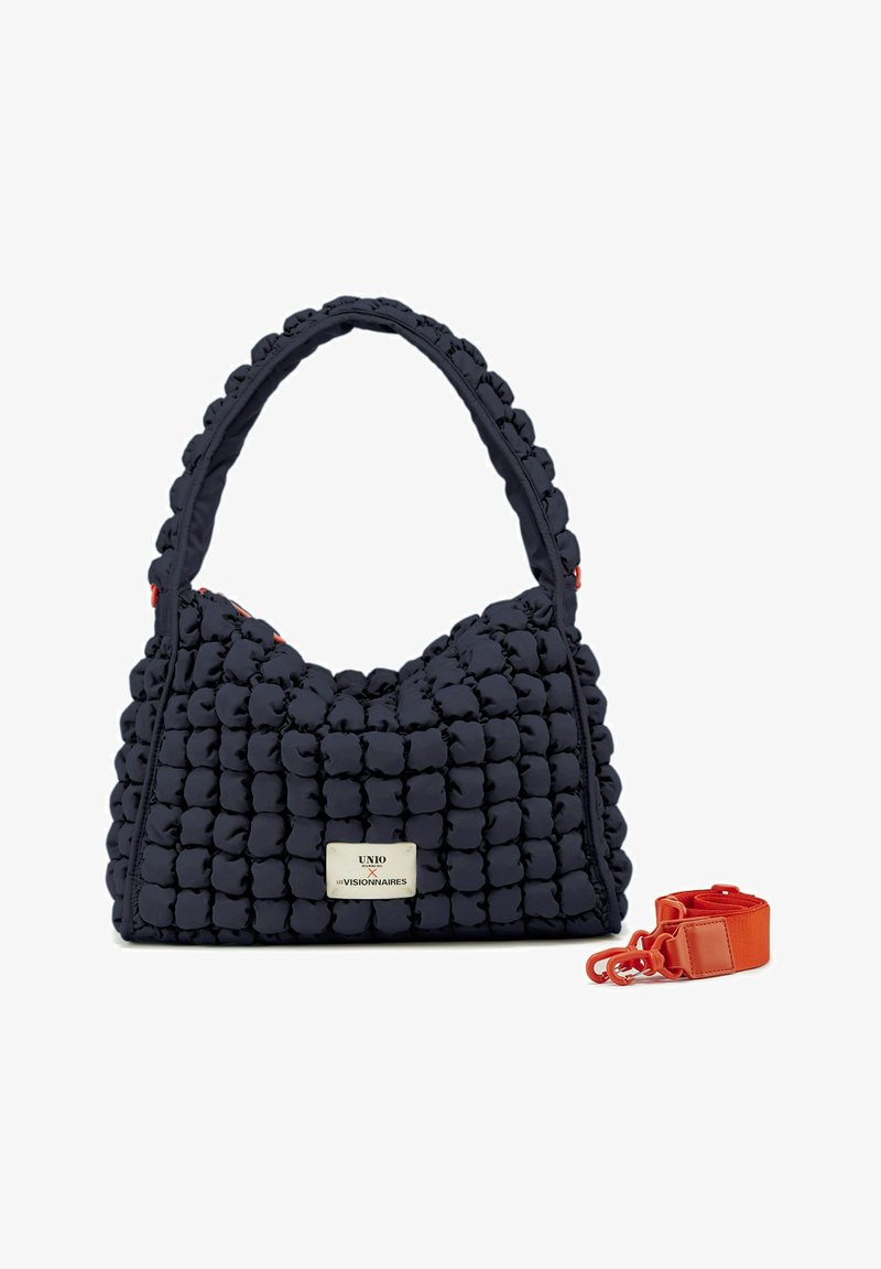 Bolso de mano acolchado de color azul marino con textura esponjosa, equipado con un asa corta y una correa naranja desmontable. Presenta una etiqueta de logo plateada.