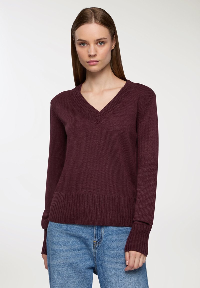 Bordowy sweter w serek z długimi rękawami, ściągaczami u dołu i na mankietach. Dzianinowa tekstura, dopasowany krój, noszony na jasnoniebieskich dżinsach.