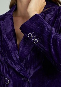 Blazer in velluto viola con design doppiopetto, rever a lancia e tre bottoni circolari decorativi sui polsini. Texture ricca visibile.