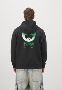 Sweat-shirt à capuche noir avec un motif circulaire vert représentant un trèfle et le texte « BOSTON EST. 1946 ». Tissu doux et coupe décontractée.