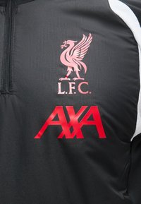 Λογότυπο της ποδοσφαιρικής ομάδας Liverpool και κόκκινο λογότυπο χορηγού AXA σε μαύρο αθλητικό μπουφάν με φερμουάρ και λευκή ρίγα.