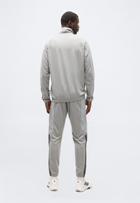 Άνδρας που στέκεται με πλάτη φορώντας ανοιχτό γκρι φόρμα με σκού ρες πλευρικές πινελιές και λευκά αθλητικά παπούτσια με μαύρες ρίγες.