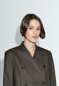 Giacca marrone doppiopetto con colletto a scialle, tessuto strutturato e due bottoni scuri. Il modello ha i capelli corti su uno sfondo chiaro.