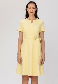 Robe jaune courte avec des manches courtes, décolleté en V, taille cintrée et détail de ceinture avec un nœud. Tissu lisse, jupe jusqu'aux genoux.