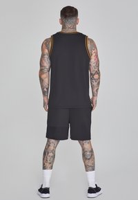 Svart ärmlös t-shirt med gula accenter, kombinerad med svarta shorts. Texturen är slät och det finns detaljerade tatueringar på benen.