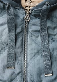 Vadderad ljusblå jacka med dragkedja, med präglade blå snören och silverfärgad hårdvara. Inre etikett med texten "HITTA PARADIS DÄR DU."