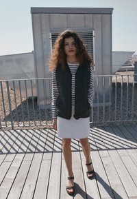 Gilet trapuntato nero sopra una maglietta a maniche lunghe bianca e nera a righe, abbinato a una gonna bianca e sandali neri con plateau, su un ponte in legno.
