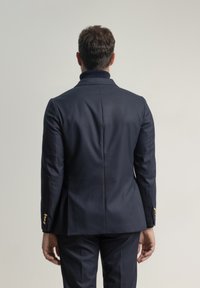 Americana de traje azul marino con un ajuste entallado, textura suave y botones dorados en los puños, con un cuello estructurado y una abertura trasera.