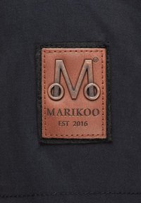 Odinio ženklo etiketė su iškilniu „M“ ir apvaliais akcentais, pritvirtinta prie tamsaus audinio fono, su užrašu „MARIKOO EST 2016“. 