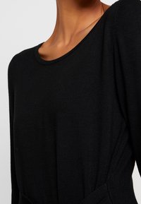 Gros plan sur une personne portant un pull en maille noir à manches longues avec un col rond et un détail de taille cintrée.