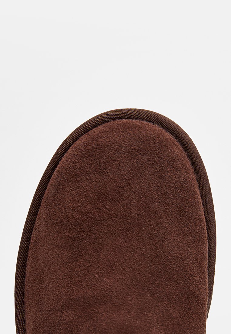 Pantoufle en cuir suédé marron avec un bout arrondi, présentant une surface texturée et une bordure en tissu marron foncé autour de la semelle.