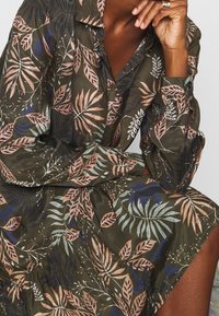 Sukienka w liście w ciemnozielonym materiale z różowymi, niebieskimi i turkusowymi akcentami; posiada kołnierz, guziki oraz zgromadzone rękawy.